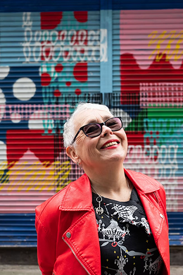 Gale laughing in front of a colourful painted wall, wearing a red leather jacket and sunglasses